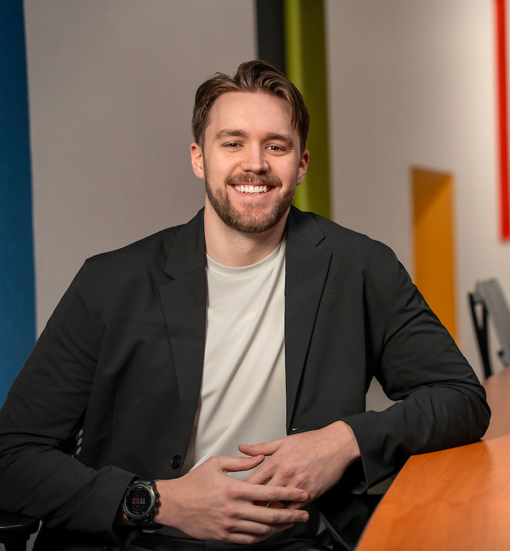 MS-FIN alum Adam Ray smiles and leans on a desk while posing for a photo